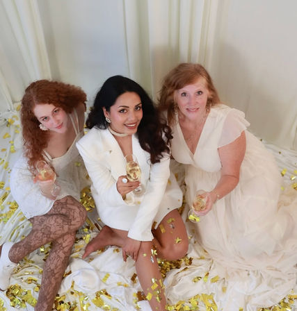 Three women in white dresses sitting on draped white fabric with gold confetti, smiling and toasting with champagne glasses in an indoor celebratory photoshoot.