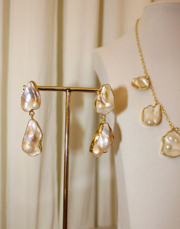 Close-up of lustrous baroque pearl drop earrings with irregular creamy pearls set in gold on a metal T-stand, with a matching gold-chain necklace on a mannequin bust against a neutral curtain backdrop