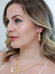 Glam close-up profile of a woman with blonde waves and glowing skin, wearing gold pearl hoop earrings and a gold necklace, with winged eyeliner and coral lips.