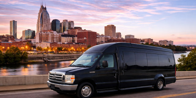 Sleek black passenger shuttle van on a riverfront road with a glowing city skyline at sunset and colorful sky reflecting on the water — urban travel and transportation scene.