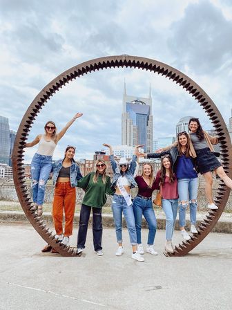 Eight friends posing and laughing inside a giant rusted circular gear sculpture with a downtown skyline backdrop, celebrating outdoors — bachelorette wearing sash and cowboy hat.