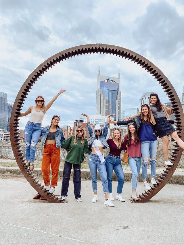 Eight friends posing and laughing inside a giant rusted circular gear sculpture with a downtown skyline backdrop, celebrating outdoors — bachelorette wearing sash and cowboy hat.
