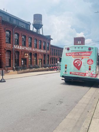 Turquoise sightseeing bus parked beside a red-brick historic factory with rooftop water tower under a cloudy sky in Nashville