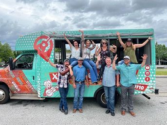 Energetic group of eight adults cheering and posing at a turquoise polka-dot food truck, some sitting on the serving ledge, in a parking lot under an overcast sky — outdoor food truck group photo.