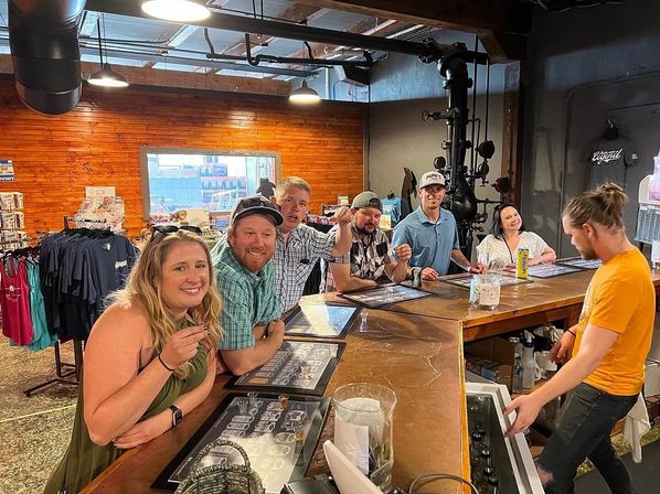Smiling group of people enjoying beer flights at a rustic industrial craft brewery taproom with wood-paneled wall, hanging lights, merch racks and a bartender serving at a long wooden tasting bar.
