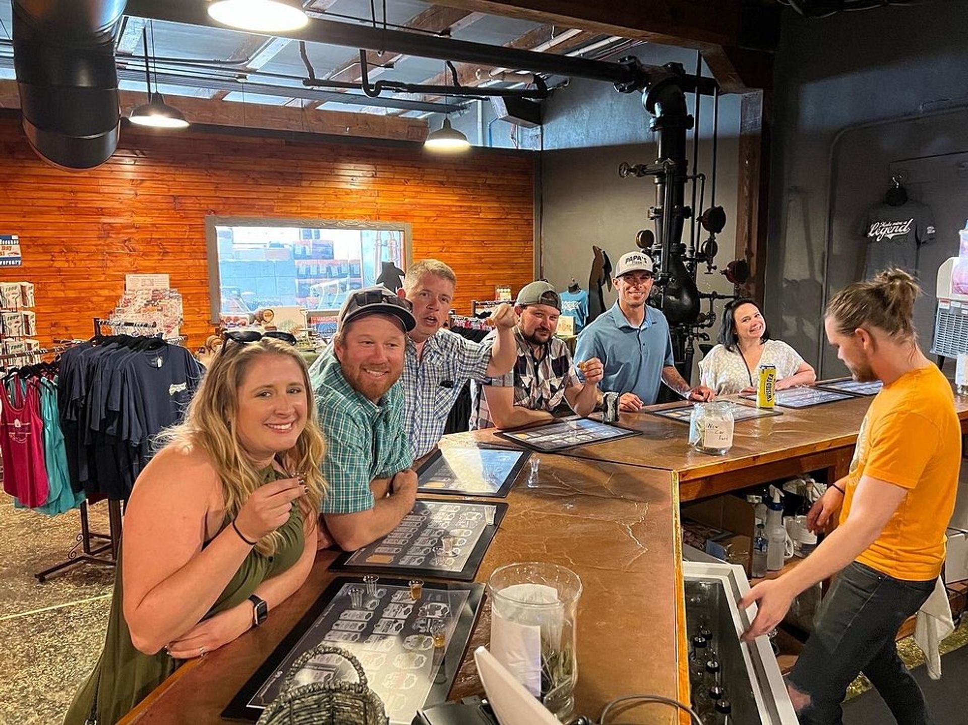 Smiling group of people enjoying beer flights at a rustic industrial craft brewery taproom with wood-paneled wall, hanging lights, merch racks and a bartender serving at a long wooden tasting bar.