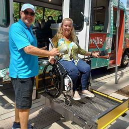 Smiling woman in a wheelchair uses a bus wheelchair lift while an attendant in a blue shirt assists beside a colorful accessible transit vehicle on a sunny city sidewalk.