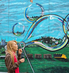 Person blowing a soap bubble in front of a vibrant outdoor mural of oversized reflective bubbles with miniature cityscapes and a train - playful urban street art