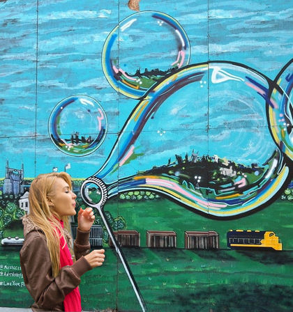 Person blowing a soap bubble in front of a vibrant outdoor mural of oversized reflective bubbles with miniature cityscapes and a train - playful urban street art