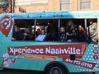 Turquoise sightseeing tour trolley in downtown Nashville with a group of smiling passengers looking out open windows against a red brick building backdrop.