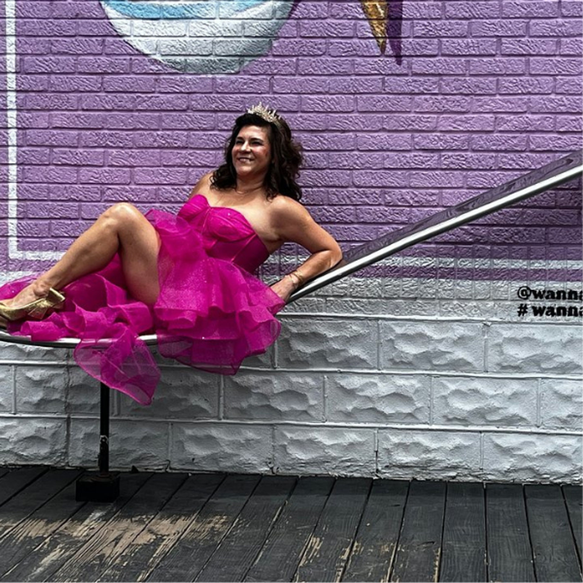 Smiling woman in a bright fuchsia tulle cocktail dress and gold tiara lounging on a metal handrail against a purple painted brick mural above a textured white base and wooden boardwalk.