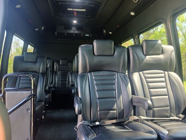 Spacious luxury passenger van interior with plush black leather captain seats, armrests, overhead emergency hatch and large windows showing green trees outside