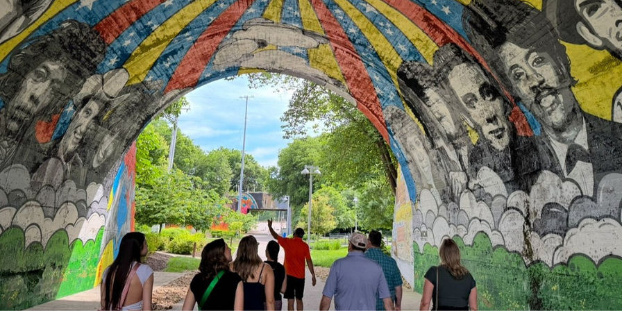 Group of people walking through a colorful mural tunnel underpass in a leafy urban park, rainbow-striped arch and large painted portraits on concrete walls