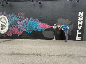 Person leans toward a painted vintage microphone on a black-brick Nashville street-art mural, colorful blue-pink-purple smoke streaming from an oversized ear