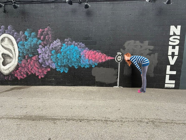 Person leans toward a painted vintage microphone on a black-brick Nashville street-art mural, colorful blue-pink-purple smoke streaming from an oversized ear