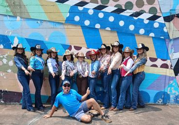 Row of women in cowboy hats and sashes posing and pointing in front of a colorful geometric street-art mural, with a man in a blue shirt reclining on the sidewalk.