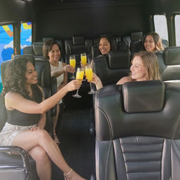 Five women clinking mimosa glasses inside a luxury party van with black leather seats — cheerful in-transit celebration.