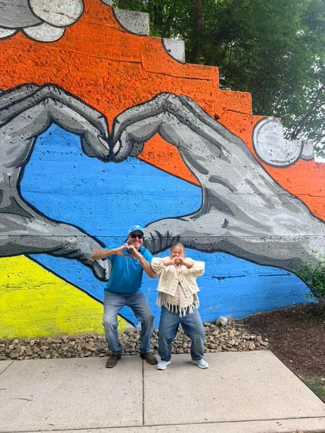 Two people making heart gestures and smiling on a sidewalk in front of a colorful outdoor street-art mural of giant hands forming a heart, with bright orange, blue and yellow background and trees above.