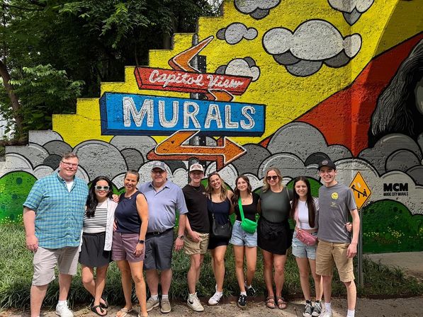 Ten people in casual summer clothes smiling and posing in front of a bright outdoor mural with clouds, bold arrows and the large painted word "MURALS".
