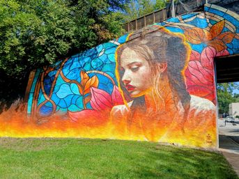 Vibrant underpass street-art mural of a dreamy female portrait with braided hair, surrounded by stained-glass-style blue and pink floral patterns and a fiery orange base above a grassy verge.