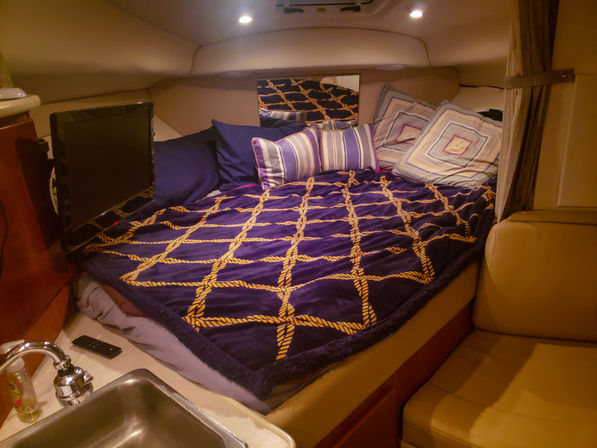 Cozy yacht cabin berth with navy blanket patterned like yellow rope, striped and geometric pillows, small TV and compact sink area