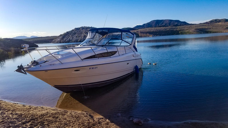 Sleek white cabin cruiser beached at a sandy lakeshore with calm blue water, rocky islets and distant desert mountains under a clear sky
