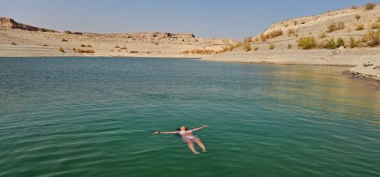 Person in a white swimsuit floating peacefully on a turquoise desert lake, framed by rocky arid shoreline and flat-topped mesas under a clear blue sky.