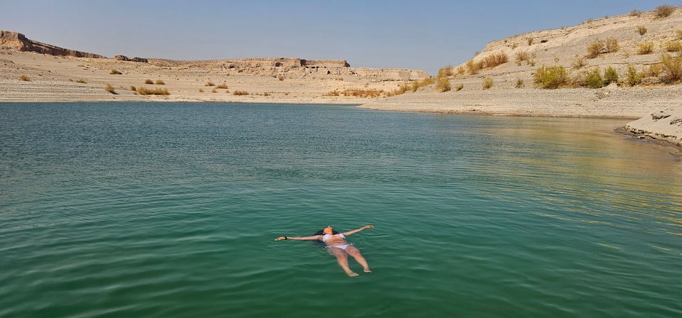 Person in a white swimsuit floating peacefully on a turquoise desert lake, framed by rocky arid shoreline and flat-topped mesas under a clear blue sky.