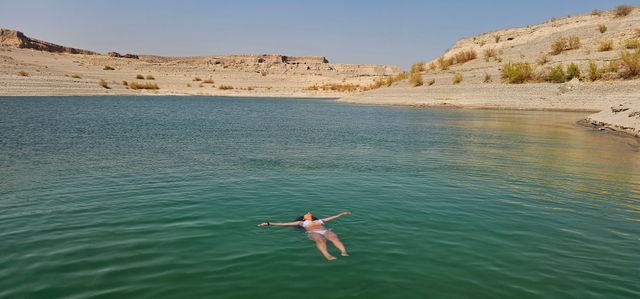 Person in a white swimsuit floating peacefully on a turquoise desert lake, framed by rocky arid shoreline and flat-topped mesas under a clear blue sky.