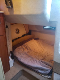 Cozy boat cabin berth with tan quilted bed, overhead bunk, small porthole window, wood cabinet and carpeted steps — compact marine sleeping area