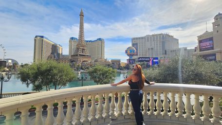 Woman on a balcony overlooking the Las Vegas Strip with an Eiffel Tower replica, lagoon, fountains and hotel skyline under a blue sky