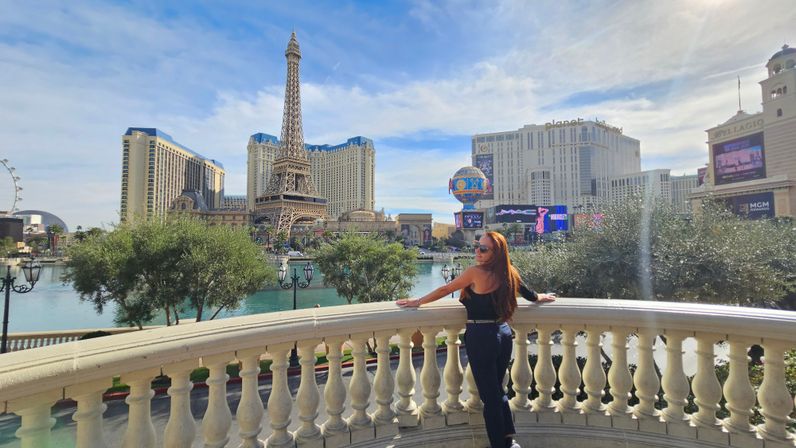 Woman on a balcony overlooking the Las Vegas Strip with an Eiffel Tower replica, lagoon, fountains and hotel skyline under a blue sky
