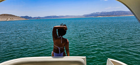 Person in a bikini at the stern of a boat looking out over turquoise lake water, desert shoreline and distant rugged mountains under a clear blue sky.