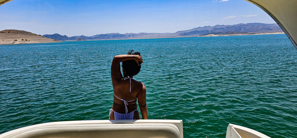 Person in a bikini at the stern of a boat looking out over turquoise lake water, desert shoreline and distant rugged mountains under a clear blue sky.