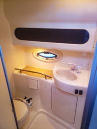 Cozy compact boat bathroom (marine head) with white sink and faucet, oval porthole window, bench with stainless rail, storage hatch and manual marine toilet.