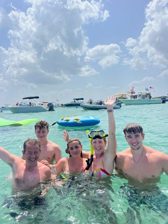 Five people smiling and waving in clear turquoise ocean water, one wearing snorkel gear, inflatable tube and anchored boats with flags in the background under a sunny, partly cloudy sky.