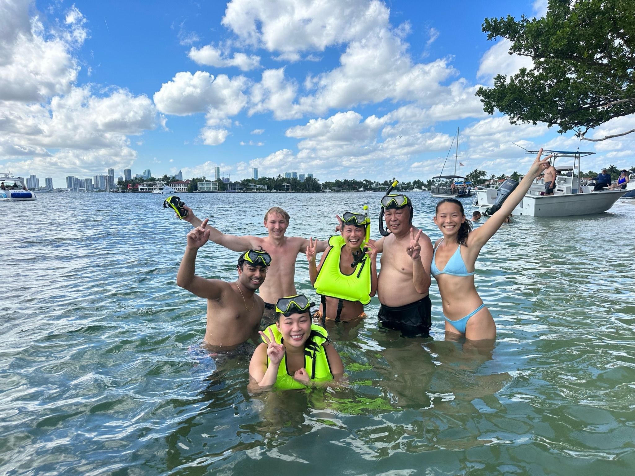 Six people in snorkel masks and bright green life vests smiling and posing in shallow bay water near boats, with an urban skyline and blue sky with puffy clouds