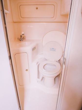 Cozy compact white boat/RV bathroom head with open toilet, foot-pedal flush, small sink faucet and a storage hatch in a fiberglass cabin