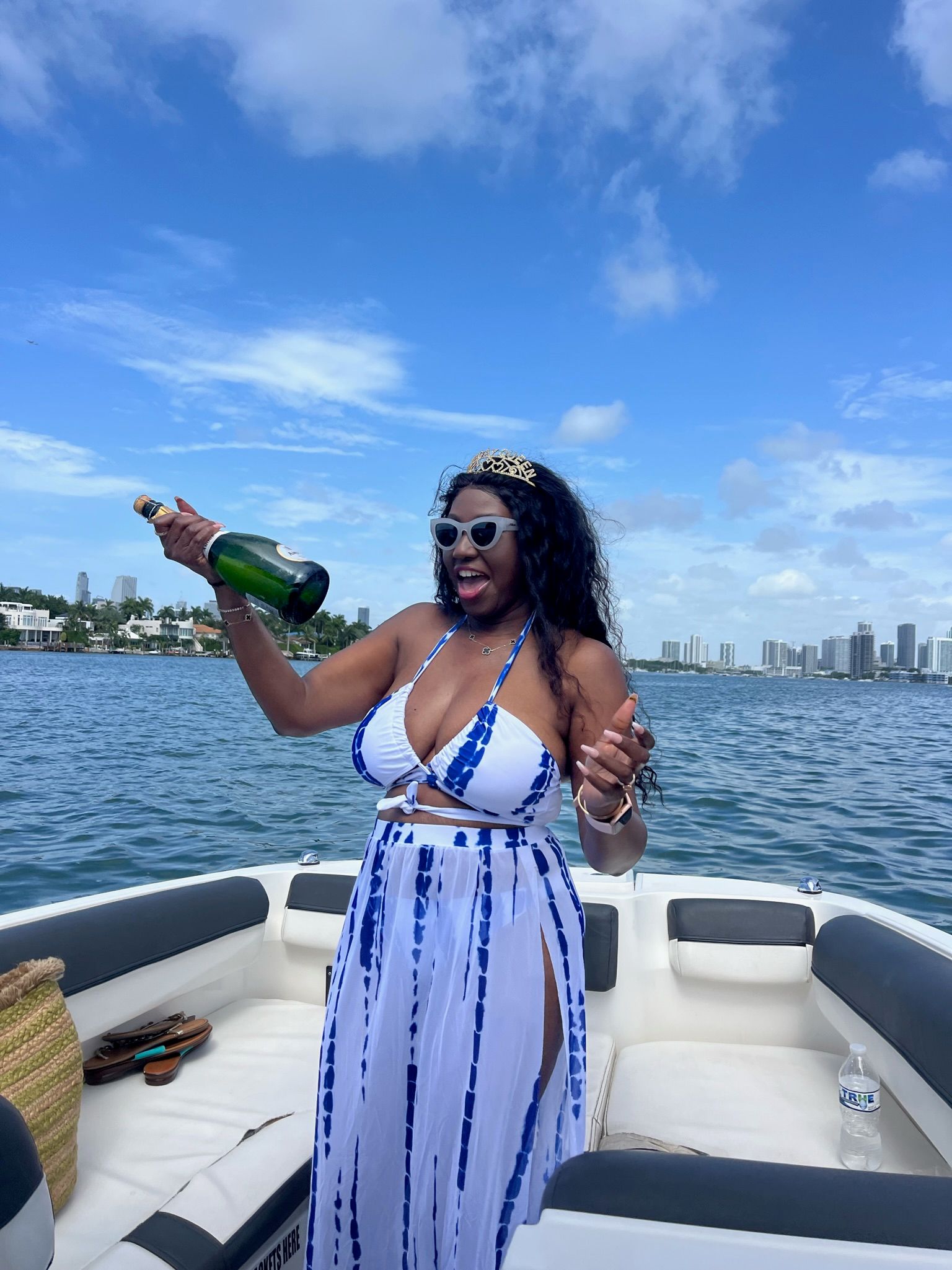 Woman in blue-and-white tie-dye bikini dress and sunglasses popping a champagne bottle on a motorboat with Miami high-rise skyline and blue sky