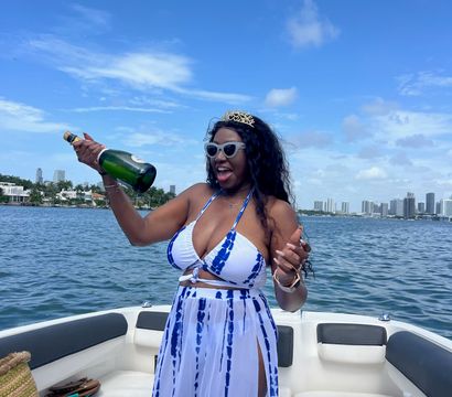 Woman in blue-and-white tie-dye bikini dress and sunglasses popping a champagne bottle on a motorboat with Miami high-rise skyline and blue sky