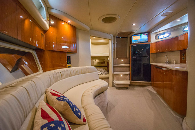 Cozy luxury yacht salon with curved cream leather sofa, patriotic throw pillows, glossy wood cabinets, compact galley with fridge and sink, and carpeted steps leading to a sleeping berth.