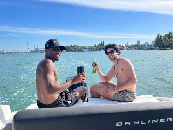 Two friends toasting with drinks on the stern of a motorboat, cruising past a palm-lined waterfront and distant city skyline on a sunny day