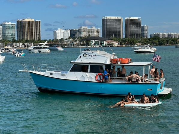 Turquoise party boat anchored in sunny coastal waters with people socializing on deck and on a paddleboard, American flag flying and high-rise waterfront skyline in the background.
