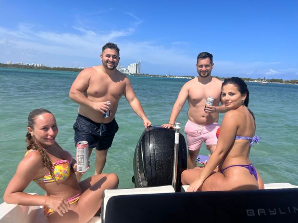 Four people in swimsuits enjoying canned drinks on a small boat in turquoise coastal bay with an outboard motor visible, urban skyline on the horizon and a clear blue sky.