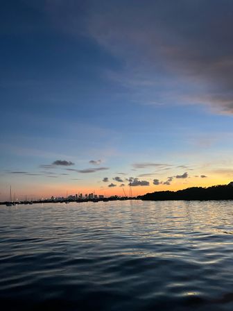 Sunset over a calm bay with a silhouetted city skyline and moored sailboats, pastel dusk sky reflecting on rippled water