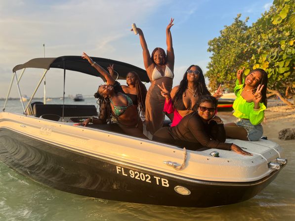Six friends in swimsuits and bright summer clothes laughing and posing on a motorboat at sunset in shallow turquoise water off a Florida beach with shoreline trees in the background.