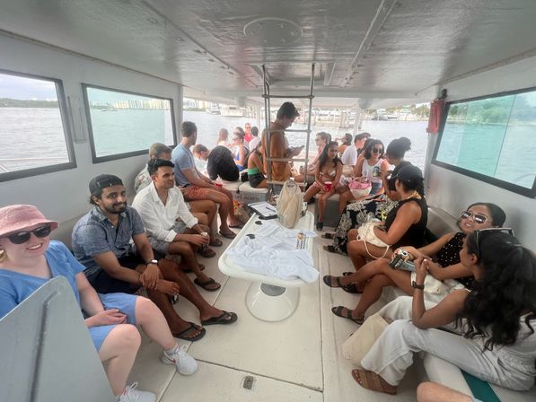 Lively group of people seated on a covered boat deck, chatting and sipping drinks with coastal water and shoreline visible through windows.