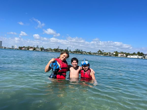 Three people in red life jackets waist-deep in clear blue bay water, smiling with snorkeling masks and fins, waterfront homes and a distant city skyline under a bright blue sky.