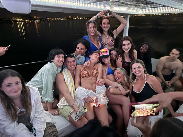 Smiling group of friends posing and taking photos at a lively nighttime boat party, in swimsuits on a lit deck with city skyline lights reflecting on the water.