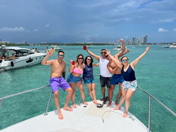 Six friends cheering on a boat bow in turquoise water near the Miami skyline, wearing swimsuits and sunglasses with red cups and yachts in the background.